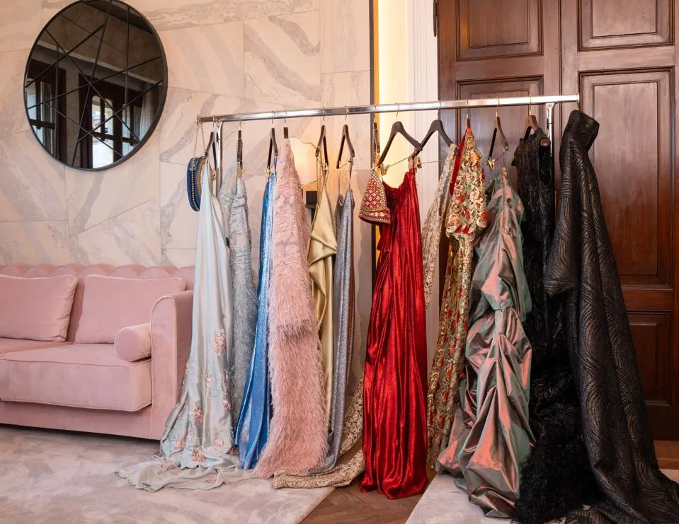 Luxury gowns, hanging from a clothes hanger inside a room. on the right is a brown door, on the left is a glimpse of the room's furniture, a pink carpet and sofa, as well as wallpaper of the same color, and above the sofa is a round mirror.