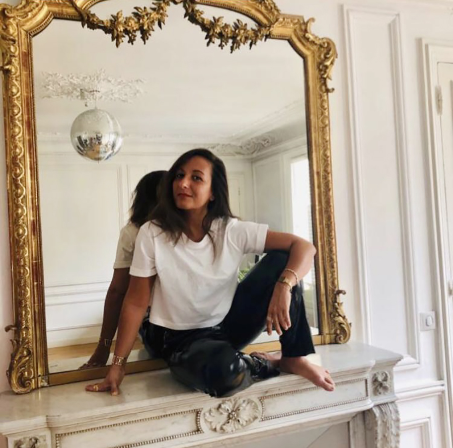 The photo depicts a smiling woman, sitting on a fireplace, inside the room of an all-white apartment. Behind her there is a big mirror with a golden frame.