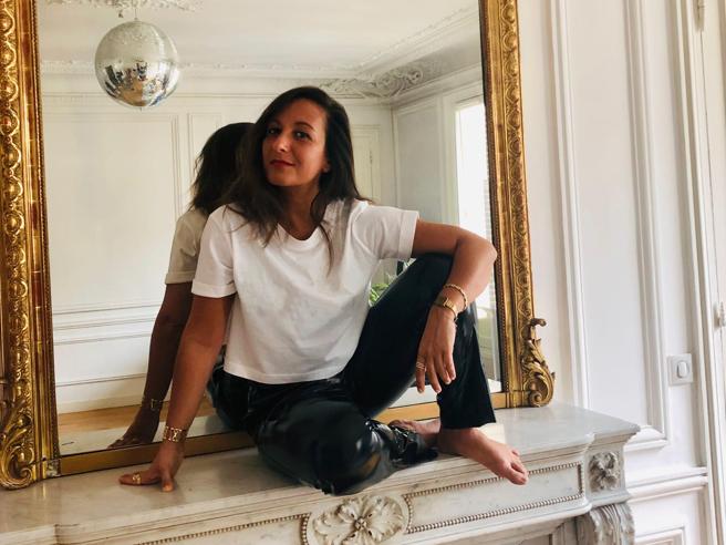 The photo depicts a smiling woman, sitting on a fireplace, inside the room of an all-white apartment. Behind her there is a big mirror with a golden frame.